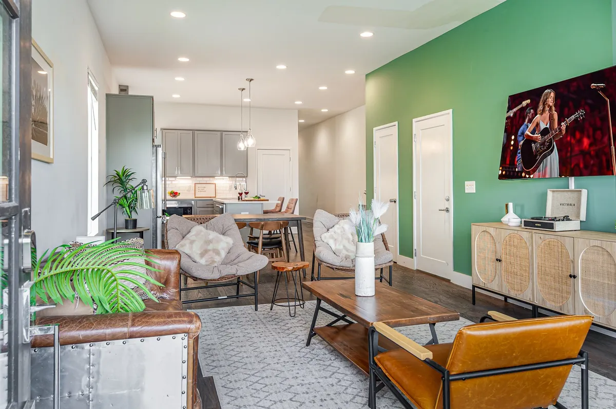 A welcoming, open-concept living area with a green accent wall, modern light fixtures, comfortable seating, a TV displaying a performance, and a dining area. The space is designed for both relaxation and group socializing, with ample natural light and an inviting ambiance.