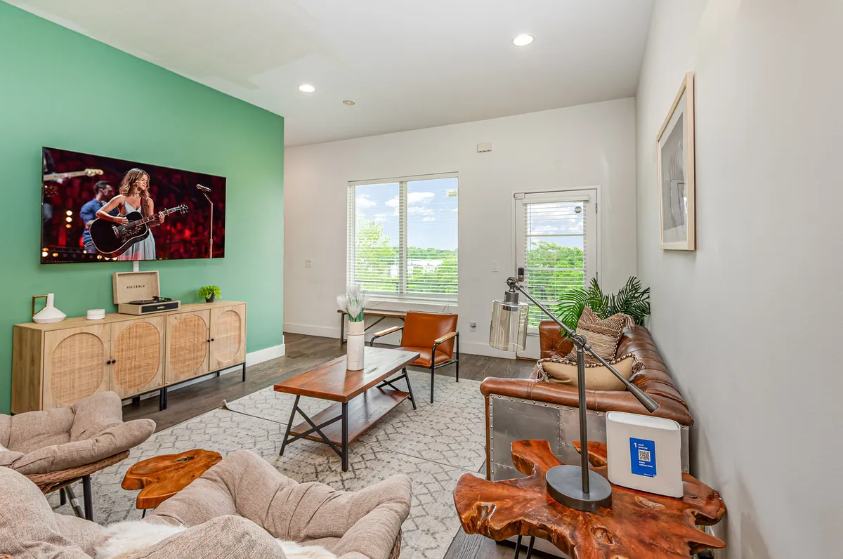 A stylish, trendy living space featuring a large TV prominently displaying a live performance, a modern sofa, an artistic coffee table, and a window with a view. The room invites entertainment and social gatherings.
