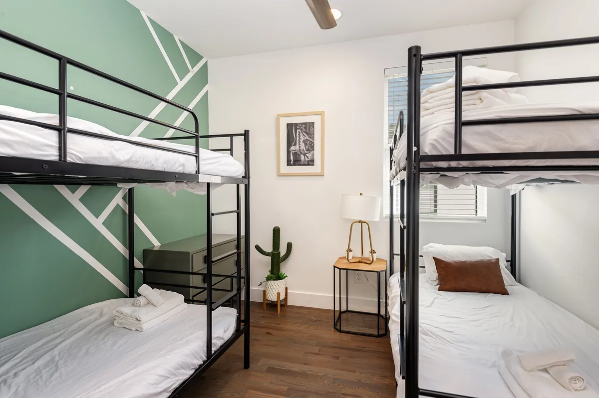 A playful bedroom with multiple bunk beds, a green accent wall, decorative cactus, lamp with side table, and framed artwork. The space feels inviting and communal, perfect for kids or groups of friends enjoying sleepovers or late-night chats.