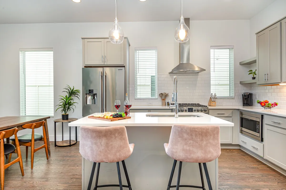 A modern, open-concept kitchen and dining area with a kitchen island, bar stools, a dining table, bright windows, and appliances. The space is designed for cooking, entertaining, and group meals, supporting both autonomy and togetherness.