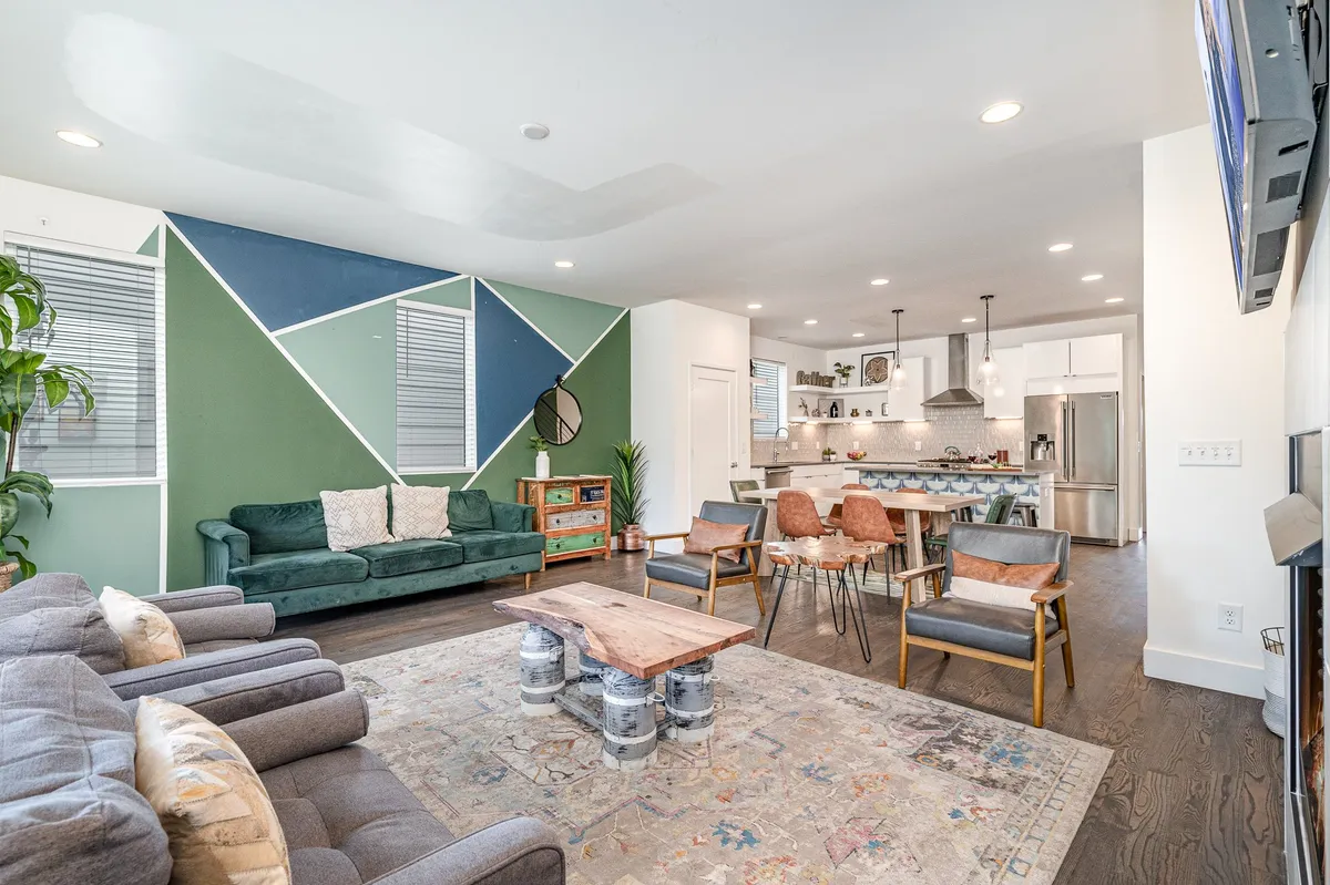 A spacious, open-concept living area featuring stylish seating, a wooden coffee table, a green accent wall with geometric patterns, and an open kitchen in the background. The room glows with natural light, inviting groups to gather, unwind, and connect.