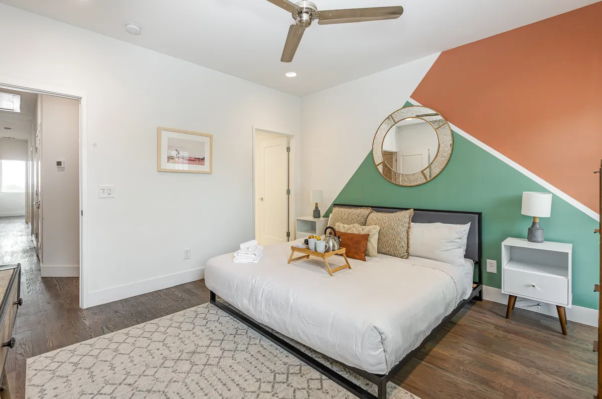 A serene bedroom featuring a comfortable bed, decorative mirror, and an accent wall, bathed in natural light. The space is styled in a cozy and modern fashion, creating an inviting atmosphere for rest.