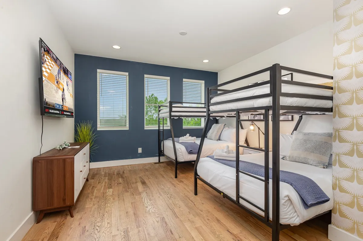 A playful and comfortable space with bunk beds, a TV atop a wooden dresser, and large windows letting in ample sunlight. The area is ideal for relaxing, watching movies, or enjoying friendly games, with enough room for socializing.