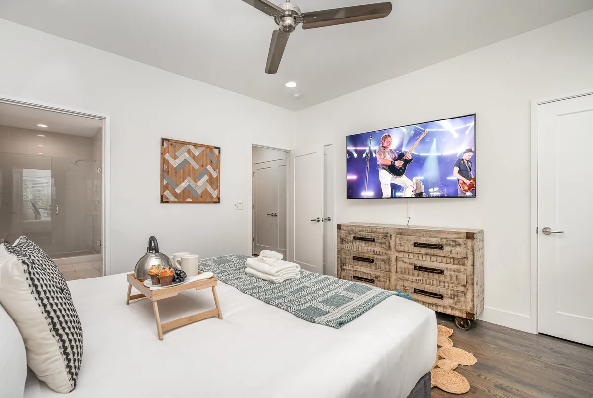 A cozy bedroom with a plush bed, decorative pillows, a wooden dresser, and a TV displaying a live band performance. Artwork on the wall adds to the lively, comfortable vibe—ideal for relaxing or enjoying music after a night out.