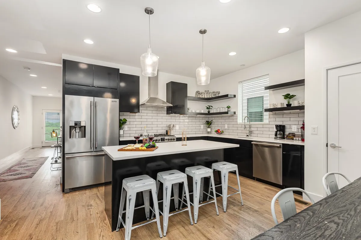 A modern kitchen featuring a spacious central island with seating, sleek black cabinetry, stainless steel appliances, a white subway tile backsplash, and decorative plants. The open layout is designed for cooking, entertaining, and gathering with friends or family.