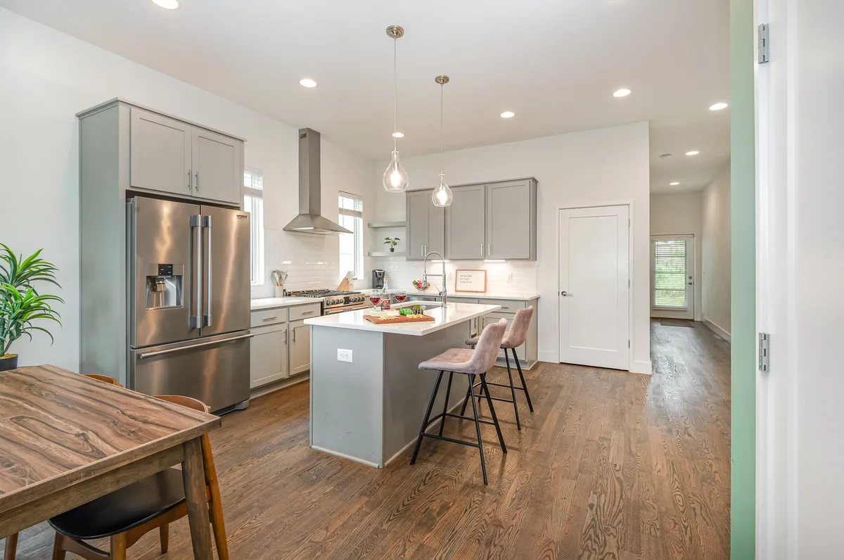 A welcoming kitchen scene with an island offering bar stool seating, a stainless steel refrigerator, visible kitchen appliances, and a wooden dining table. The setting is styled for both casual dining and entertaining.